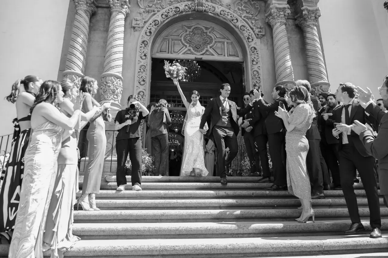 Los novios bajan las escaleras de la iglesia entre dos filas de invitados que aplauden, en blanco y negro