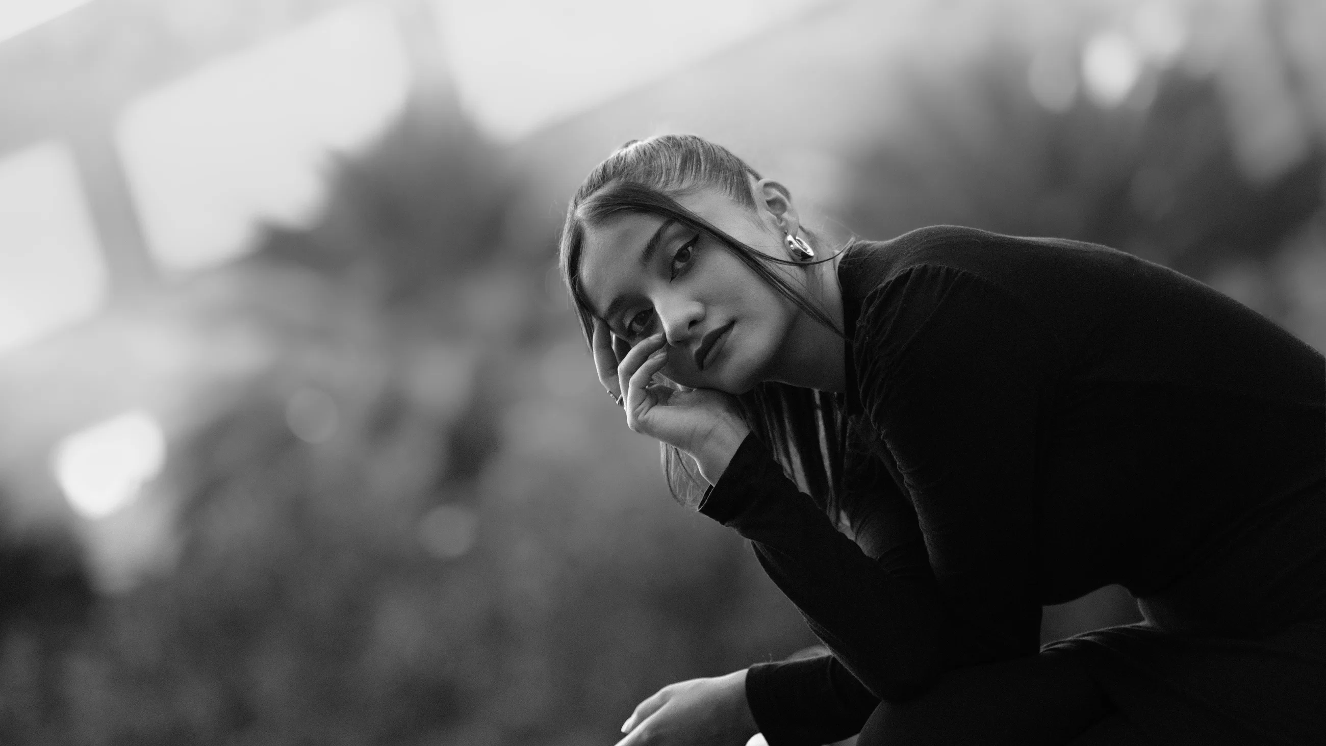 Retrato en blanco y negro de una mujer recargada en su mano, mirando a cámara, con el fondo claro completamente desenfocado detrás