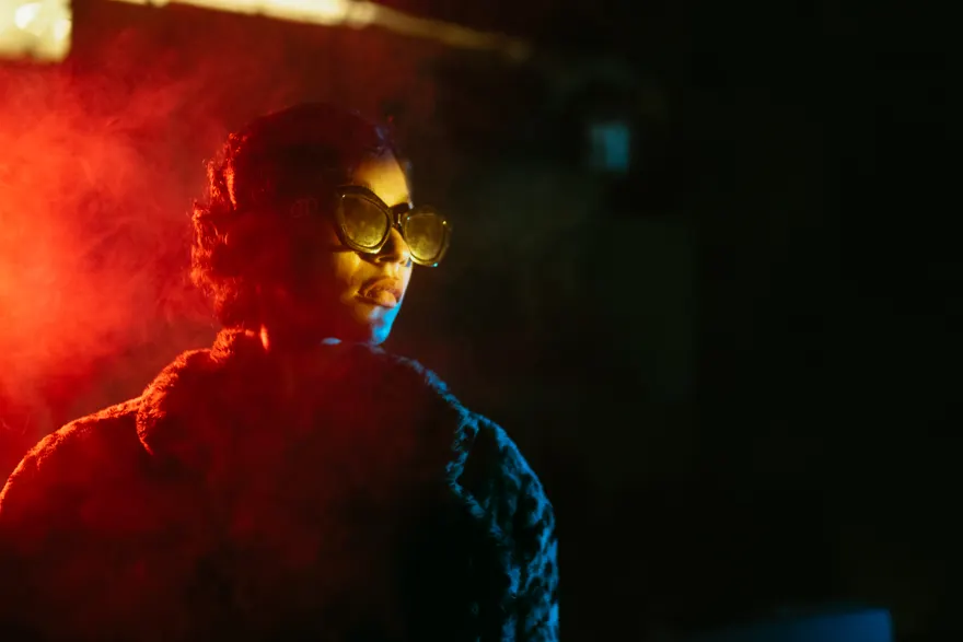 Retrato de noche volteando sobre el hombro con lentes oscuros, envuelto en humo rojo por la izquierda y con un filo de luz azul en el hombro