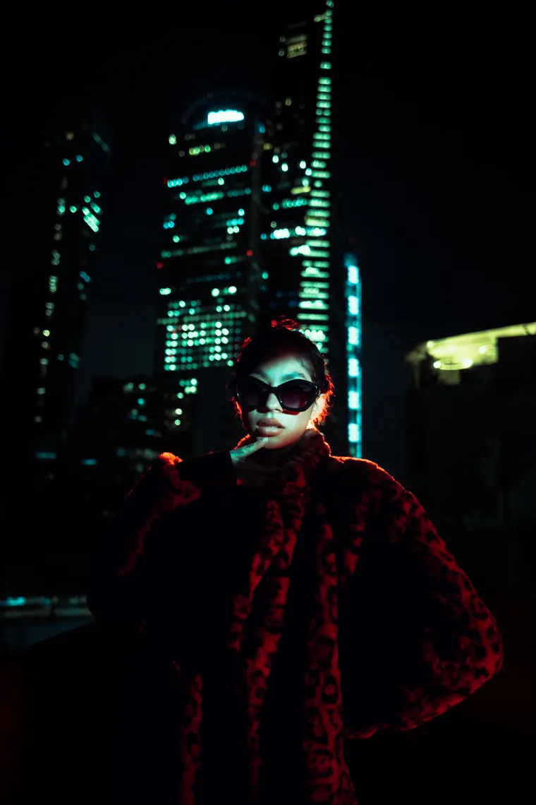Una mujer con lentes oscuros y abrigo de peluche rojo, de frente, con las torres de oficinas encendidas de noche detrás