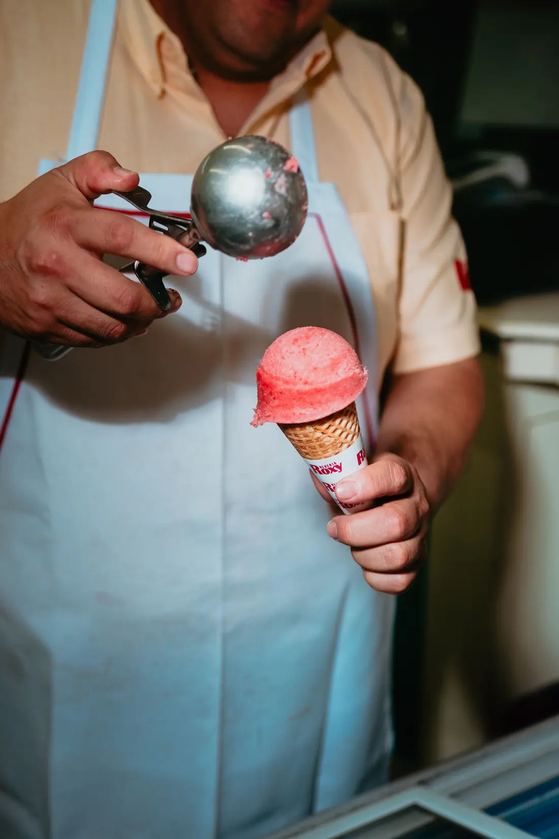 El heladero, con mandil blanco y cuchara de bola en la mano, sirve un cono de fresa