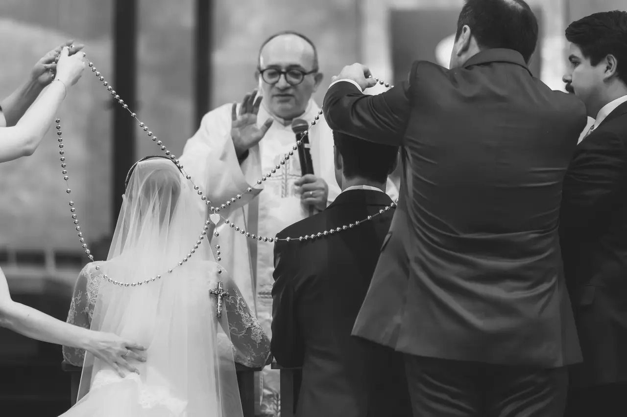 Cuatro manos colocan el lazo sobre los hombros de los novios mientras el sacerdote bendice, en blanco y negro