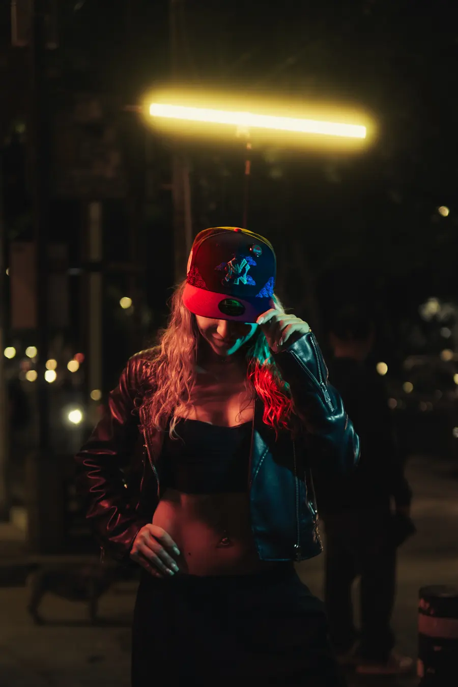 Una mujer con gorra y chamarra, de pie en la banqueta, luz naranja de la avenida