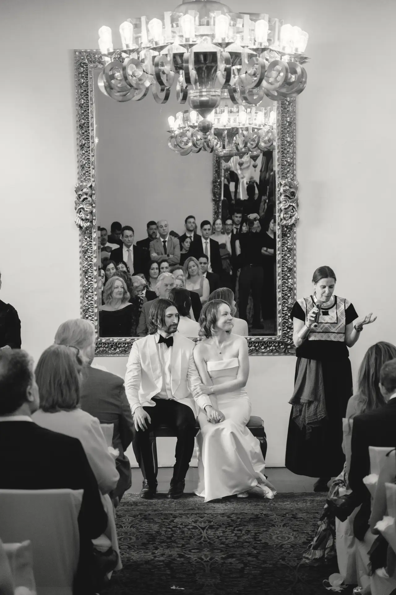 Los novios sentados de frente a la sala, con todos los invitados reflejados en el espejo que tienen detrás, en blanco y negro