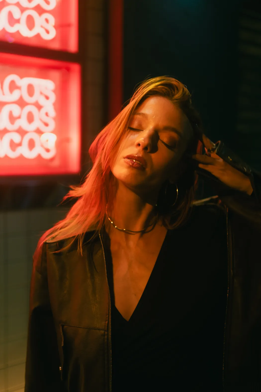 Retrato de noche con los ojos cerrados y la cabeza echada atrás, el pelo y la cara encendidos de naranja por los letreros de neón rojos que se leen detrás
