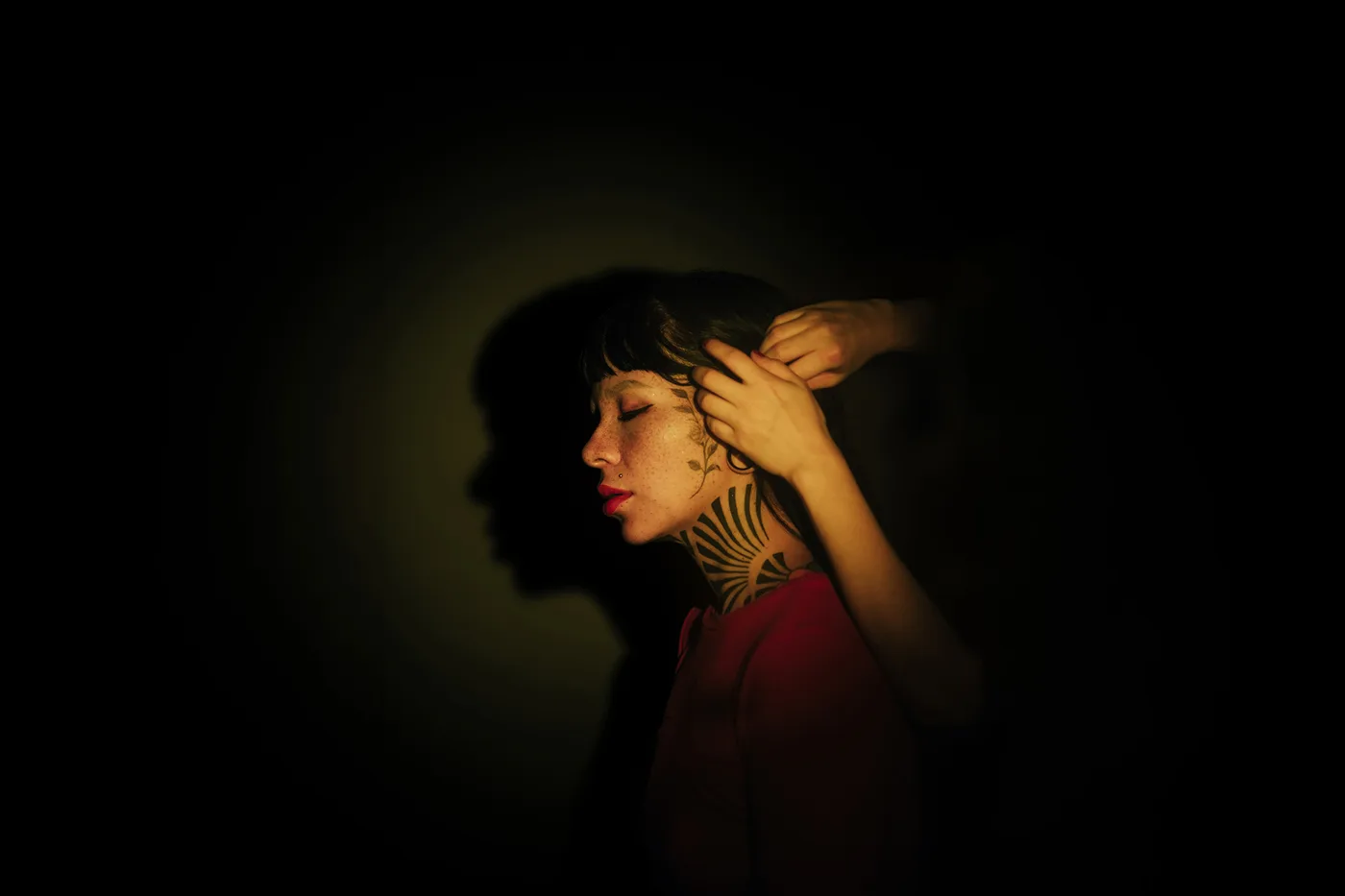 Una mujer de perfil con los ojos cerrados y la mano en el pelo, iluminada por un halo verde amarillo que proyecta su sombra enorme sobre la pared de atrás
