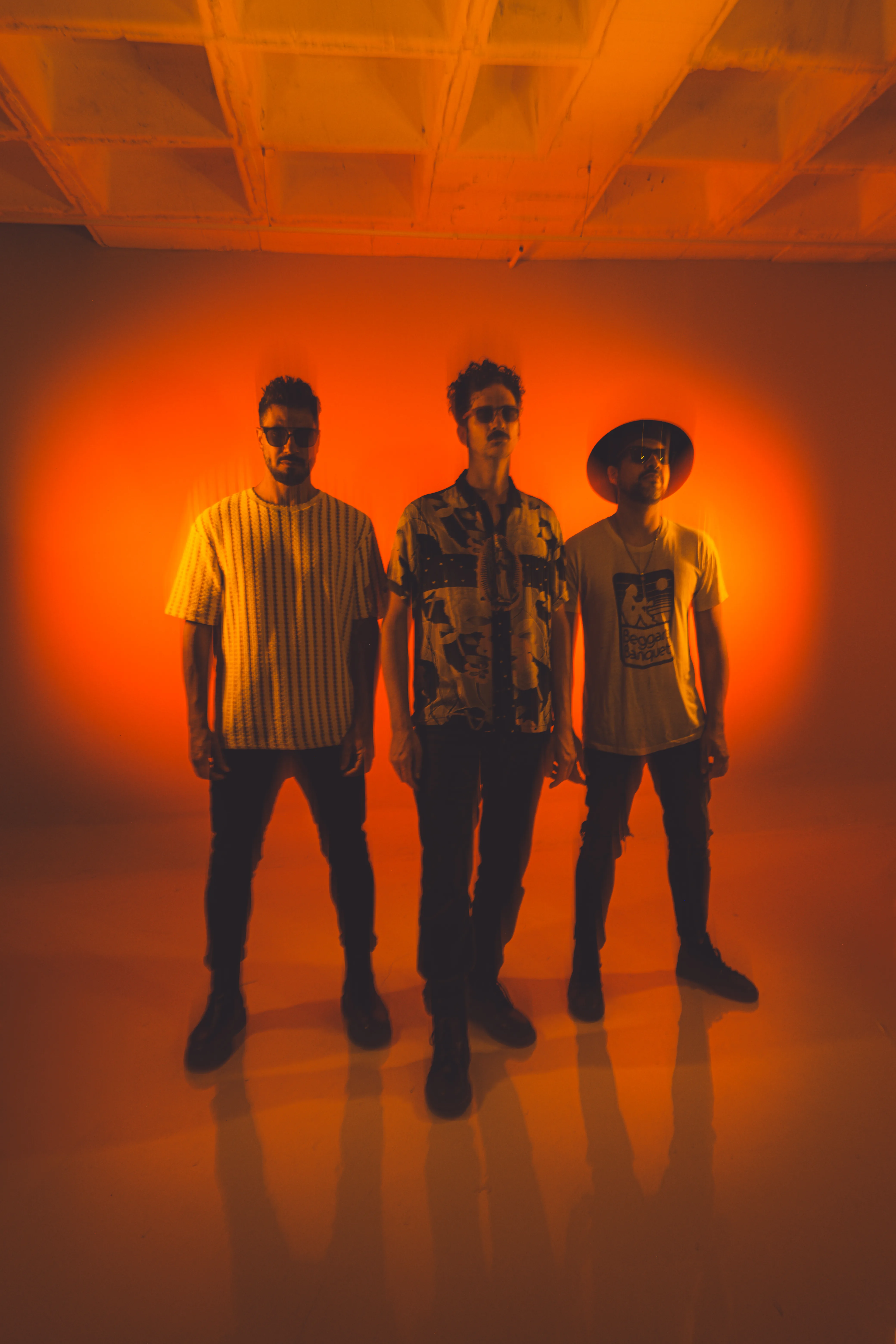 Los tres músicos de la banda, de pie ante una pared de luz naranja