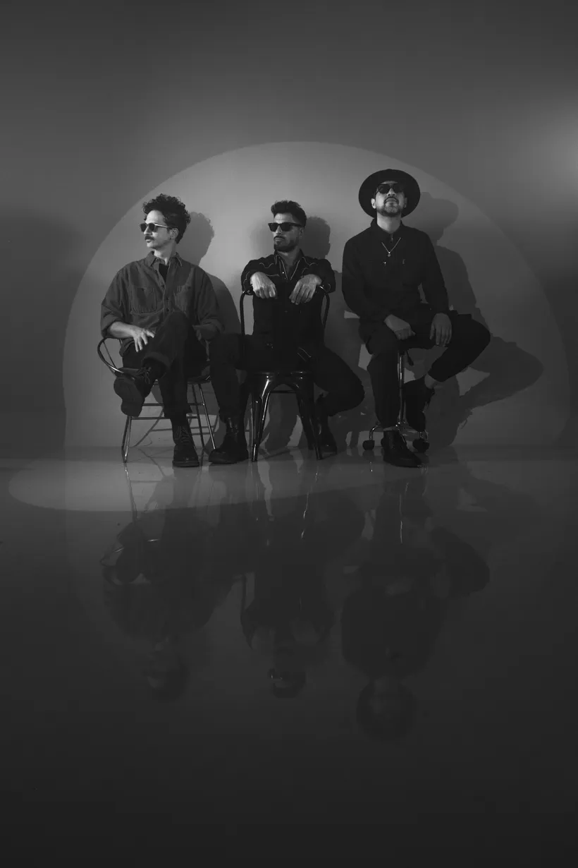 Tres hombres sentados en fila con lentes oscuros, uno de sombrero, en blanco y negro sobre un piso pulido que los refleja