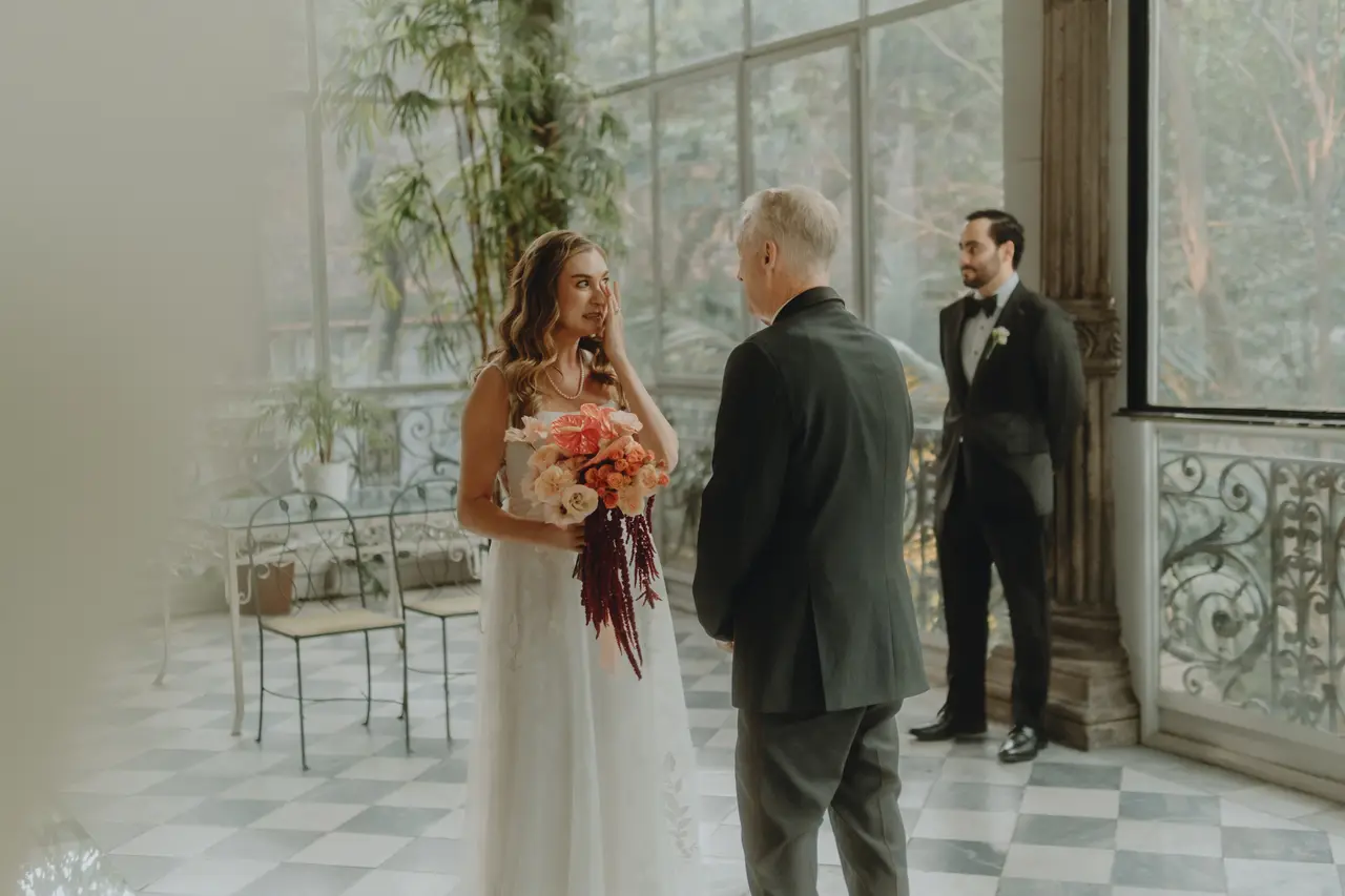 La novia se seca una lágrima junto a su padre mientras el novio la espera al fondo de la galería